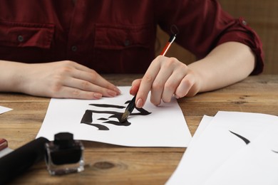 Photo of Calligraphy. Woman with brush and inkwell writing hieroglyphs on paper at wooden table, closeup