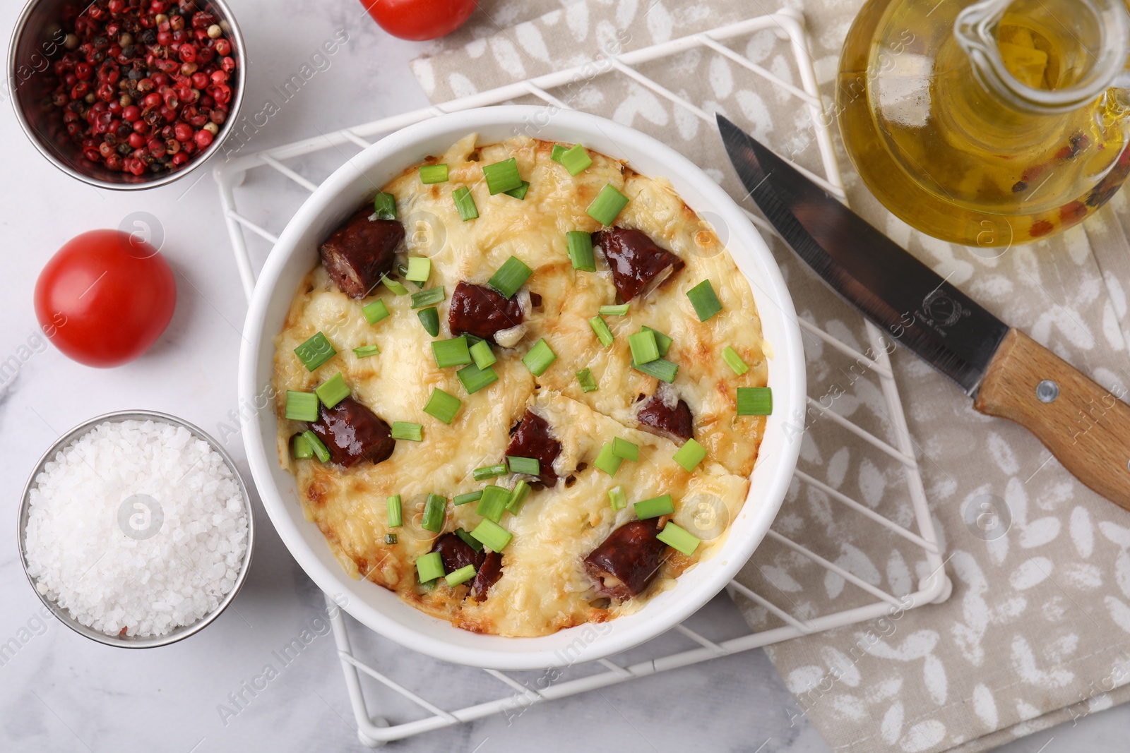 Photo of Tasty sausage casserole in baking dish, knife and ingredients on white marble table, flat lay