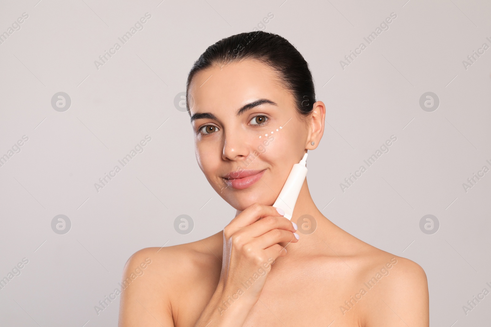 Photo of Beautiful young woman with cream on skin under eye against light grey background