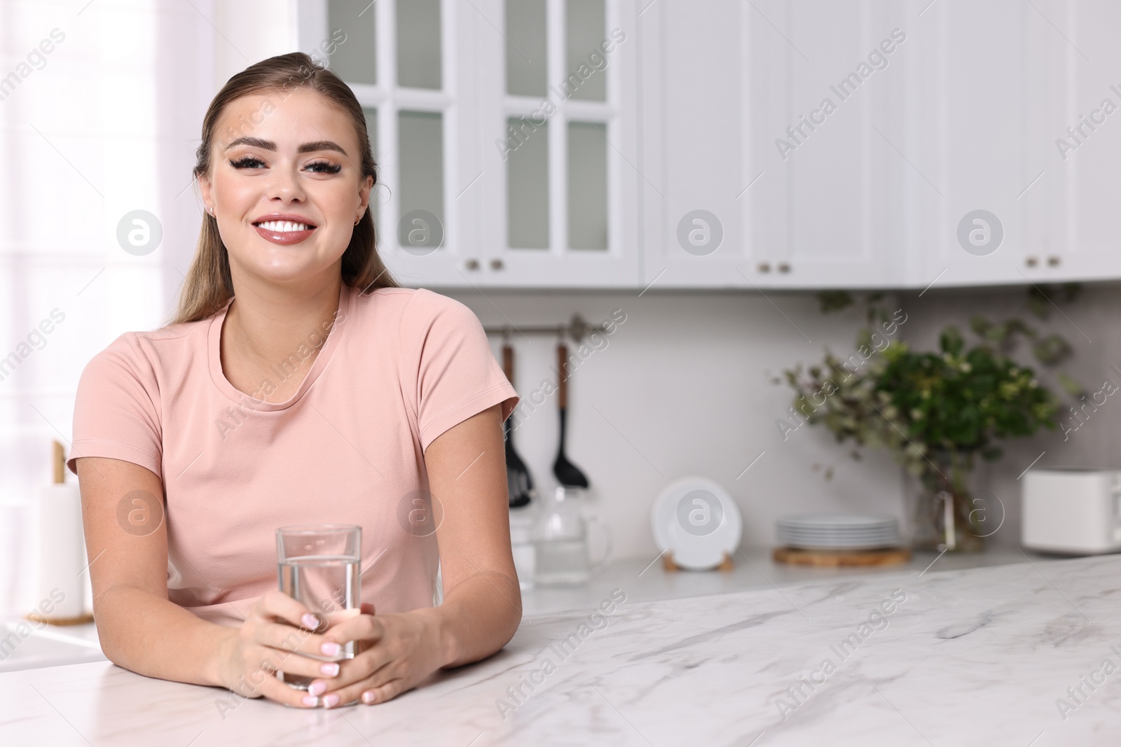 Photo of Happy woman with glass of fresh water at white marble table in kitchen, space for text