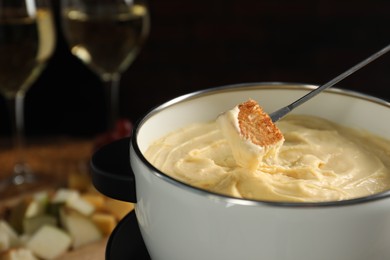 Dipping piece of bread into fondue pot with melted cheese at table, closeup