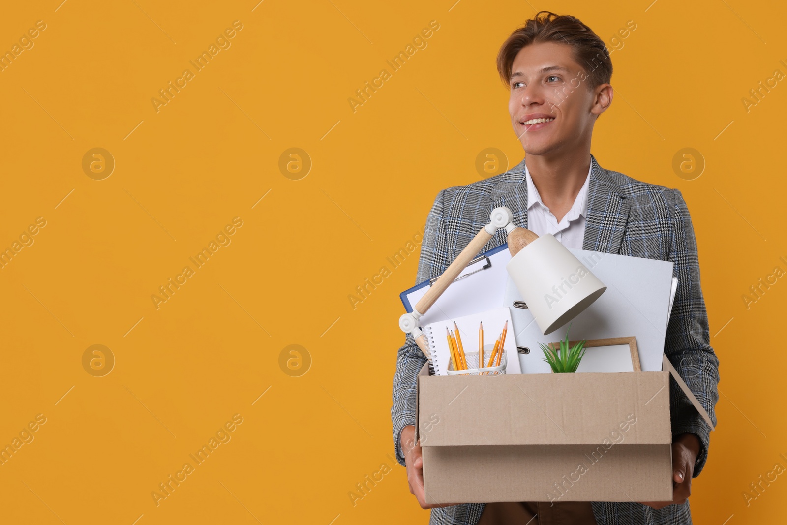 Photo of Happy unemployed young man with box of personal office belongings on orange background. Space for text