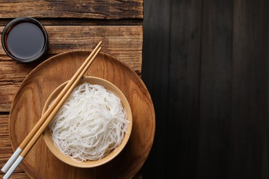 Photo of Bowl with cooked rice noodles, soy sauce and chopsticks on wooden table, flat lay. Space for text