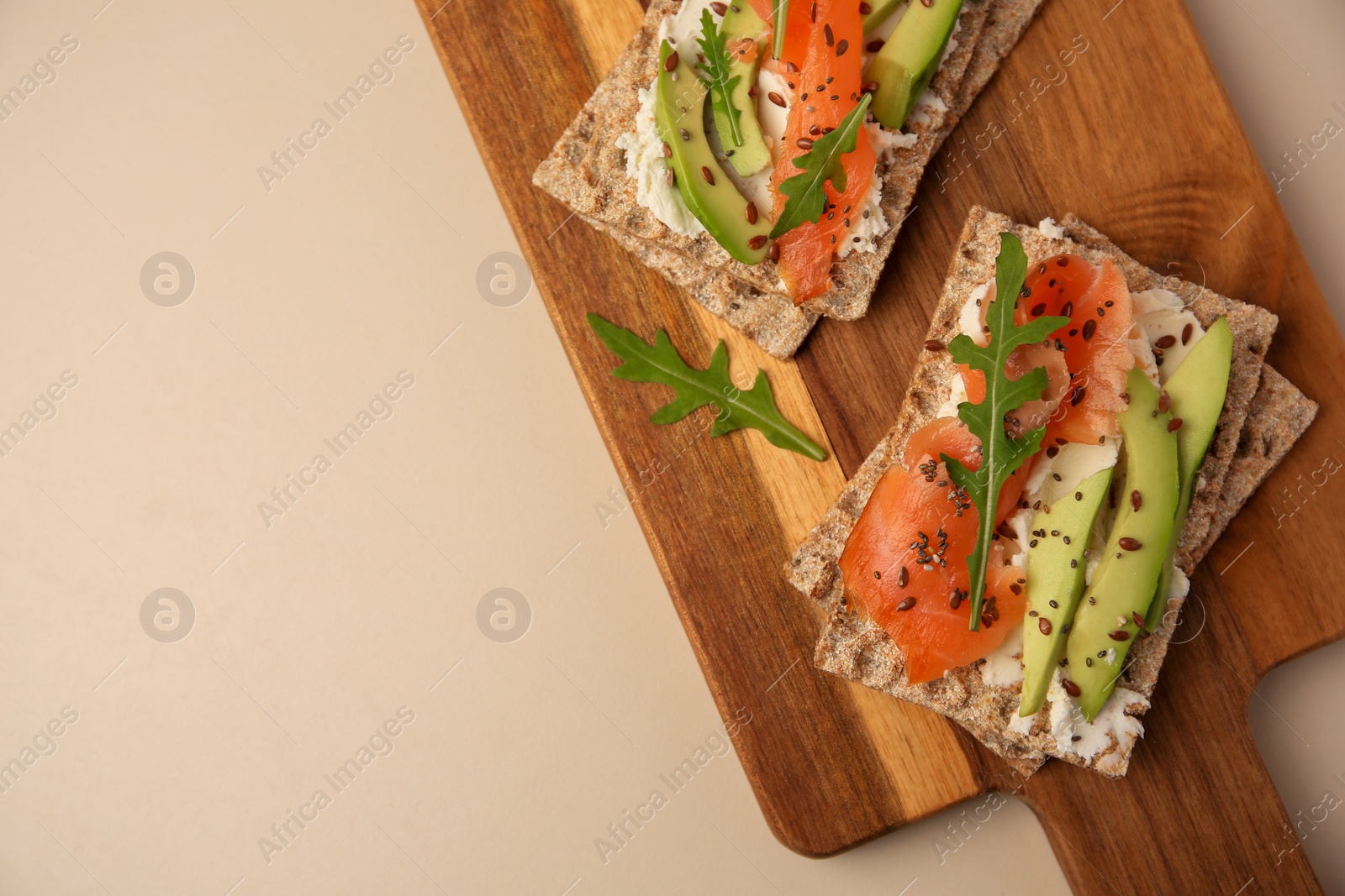 Photo of Fresh crunchy crispbreads with cream cheese, salmon, avocado and arugula on beige table, top view. Space for text
