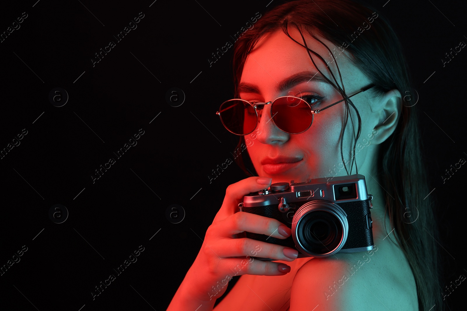 Photo of Beautiful woman with sunglasses and vintage camera posing in neon lights against black background. Space for text