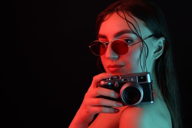 Beautiful woman with sunglasses and vintage camera posing in neon lights against black background. Space for text