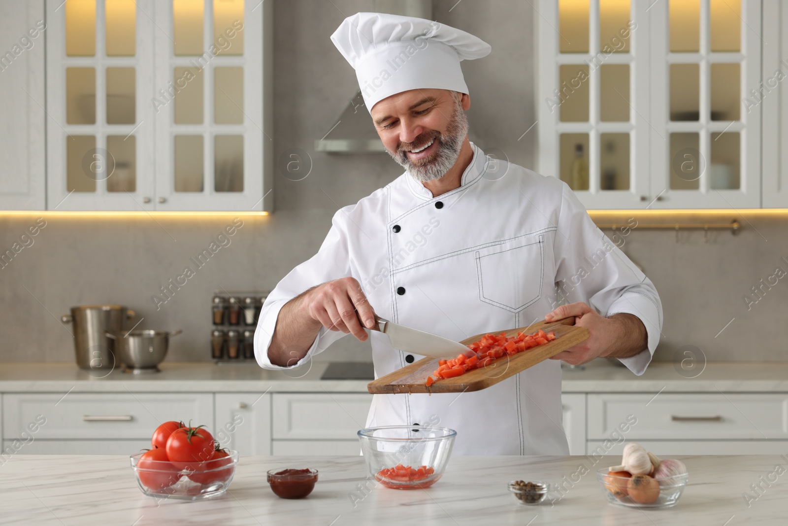 Photo of Professional chef putting cut tomatoes into bowl at white marble table indoors