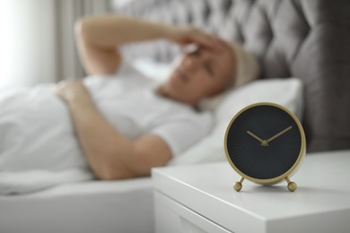 Alarm clock on nightstand with mature woman suffering from headache on background