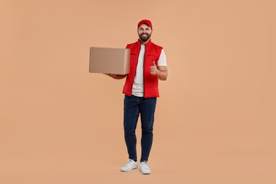 Photo of Happy young courier with parcel showing thumb up on light brown background