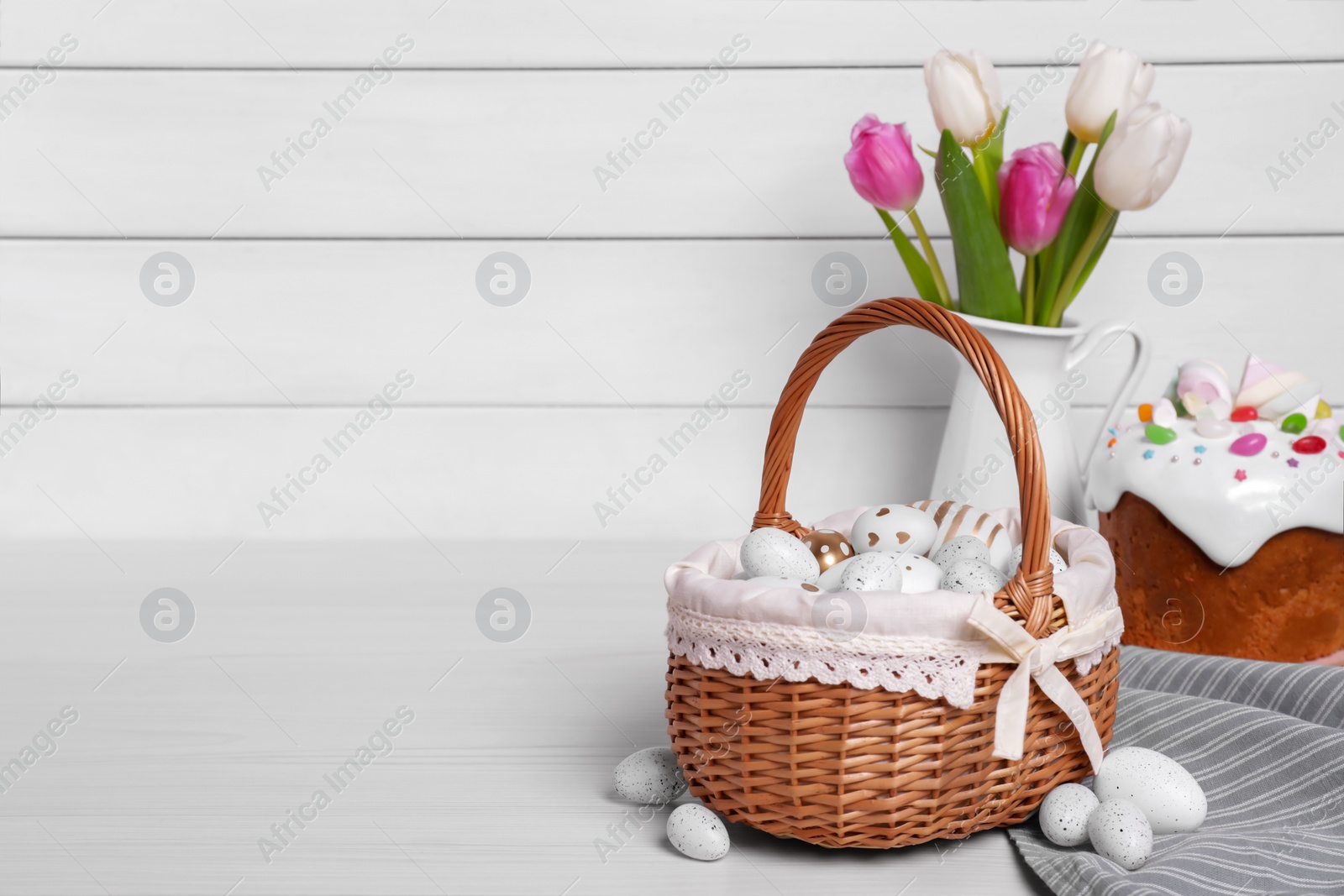 Photo of Wicker basket with festively decorated eggs, beautiful tulips and traditional Easter cake on white wooden table. Space for text