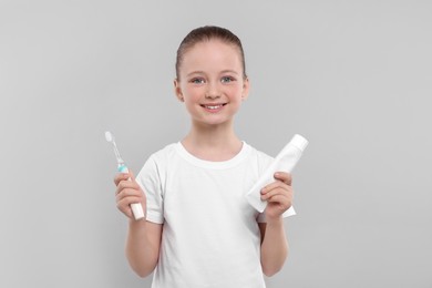 Happy girl holding electric toothbrush and tube of toothpaste on light grey background