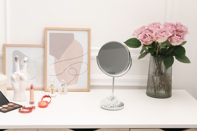 Photo of Mirror, cosmetic products, perfumes and vase with pink roses on white dressing table in makeup room