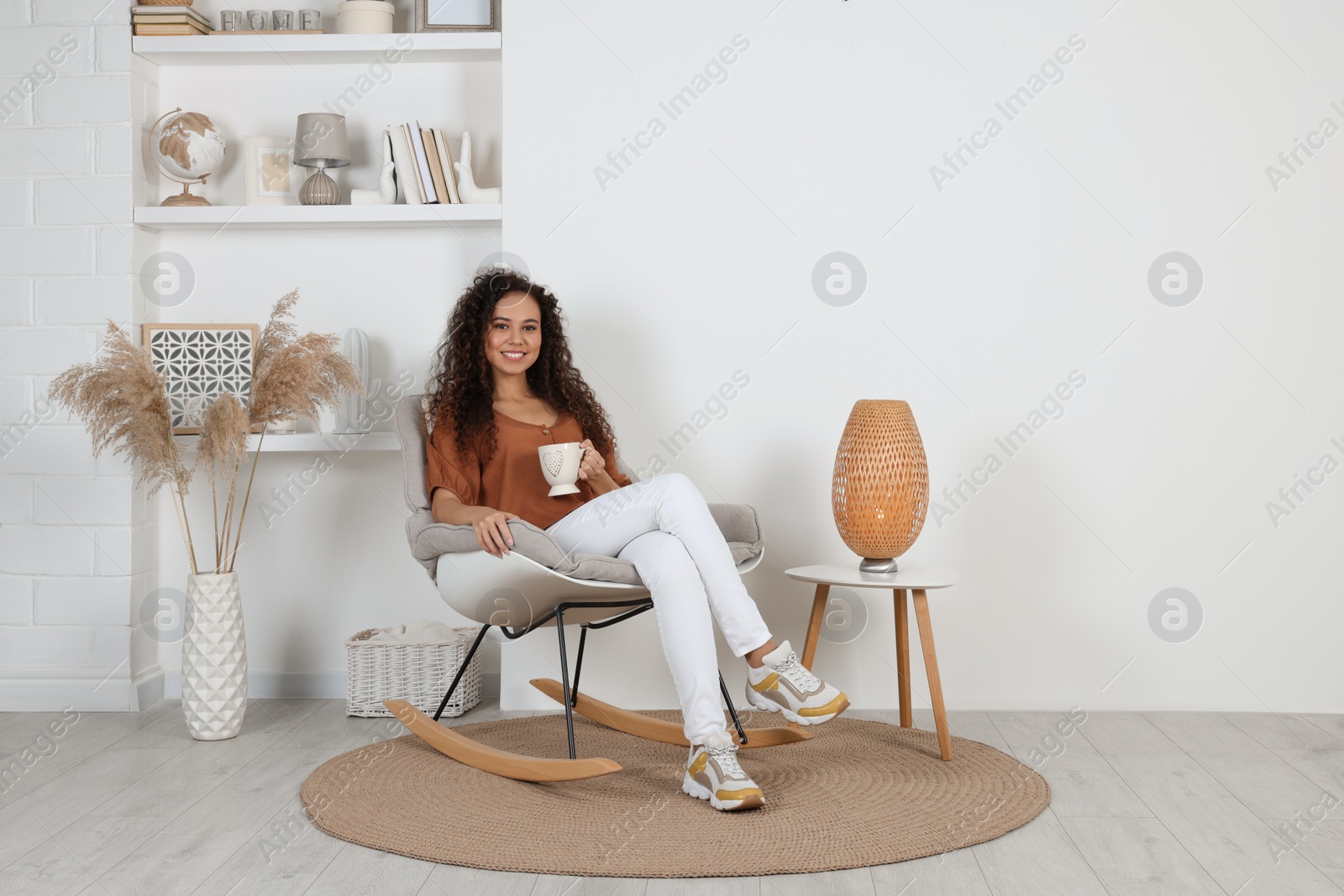 Photo of Young African-American woman with cup of drink sitting in rocking chair at home