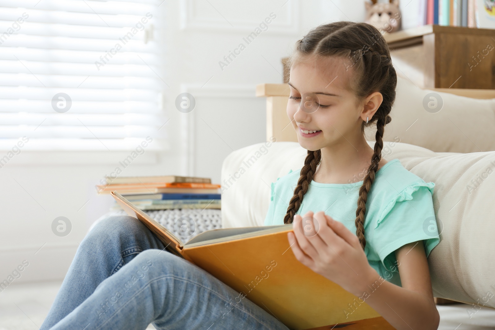 Photo of Cute little girl reading book near sofa at home