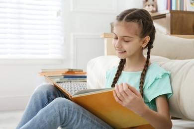 Photo of Cute little girl reading book near sofa at home