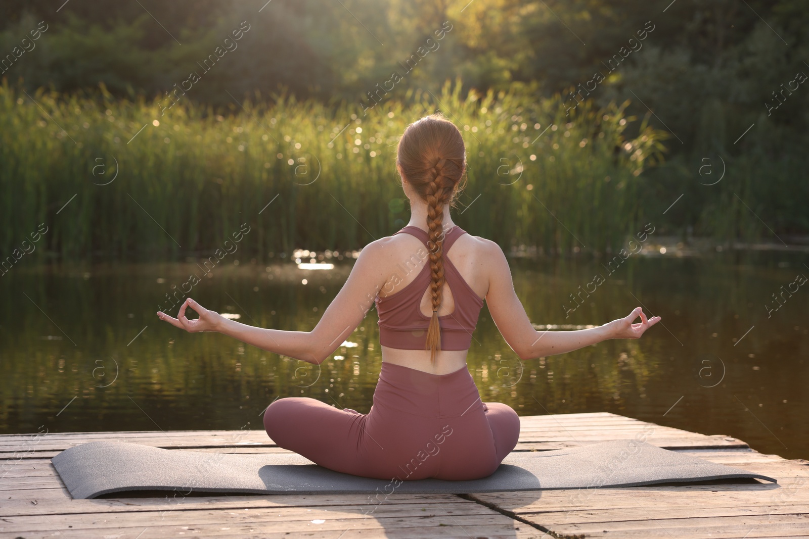 Photo of Woman practicing Padmasana on yoga mat on wooden pier near pond, back view. Lotus pose