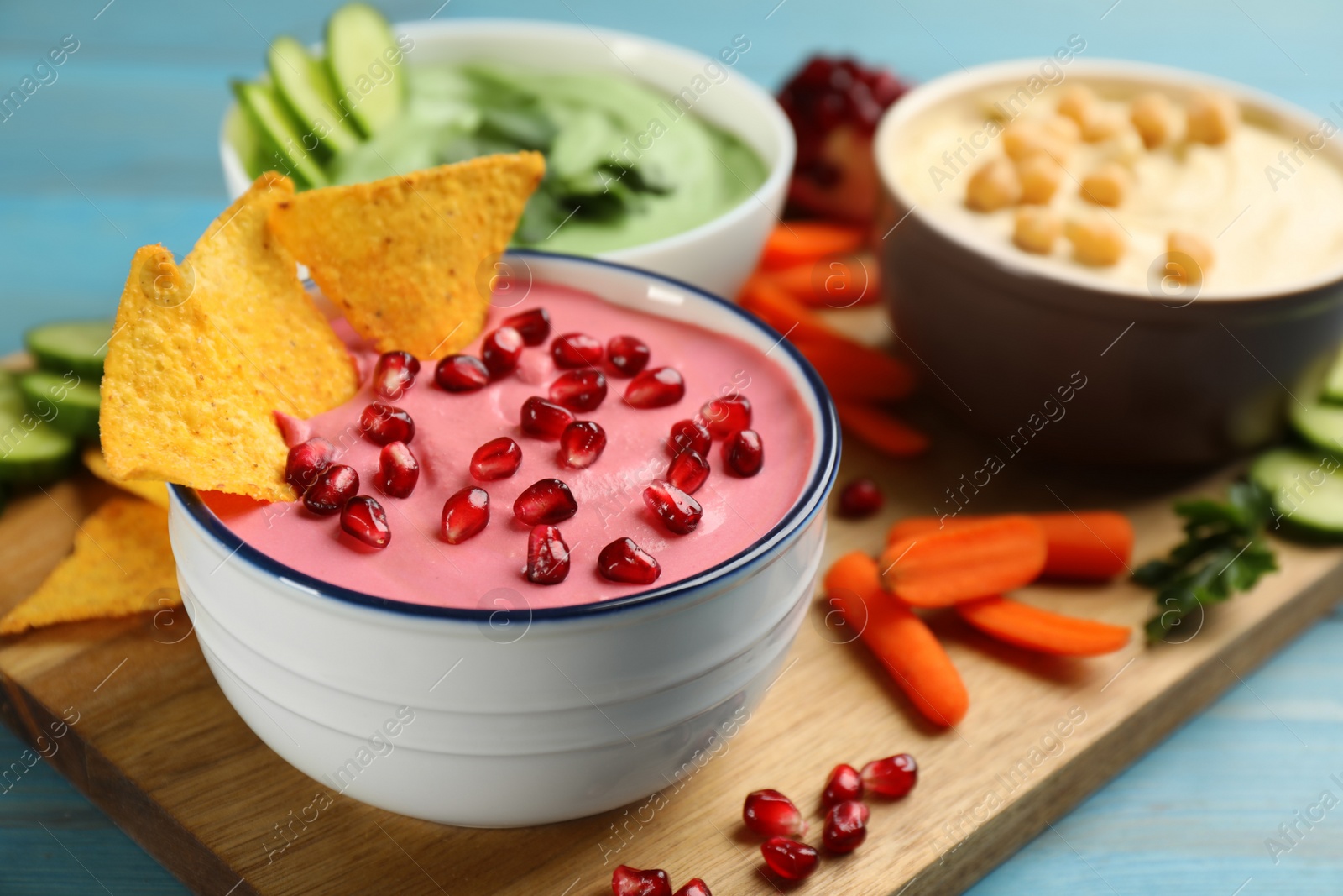 Photo of Bowl of delicious pink hummus with tortilla chips and pomegranate seeds on light blue wooden table