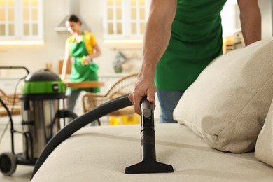 Photo of Professional janitor in uniform vacuuming sofa indoors, closeup