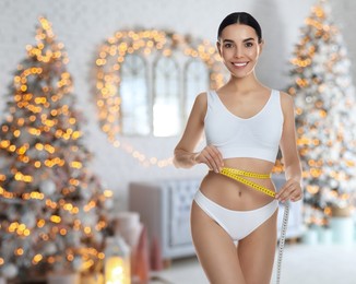 Image of Young woman measuring waist with tape in room decorated for Christmas