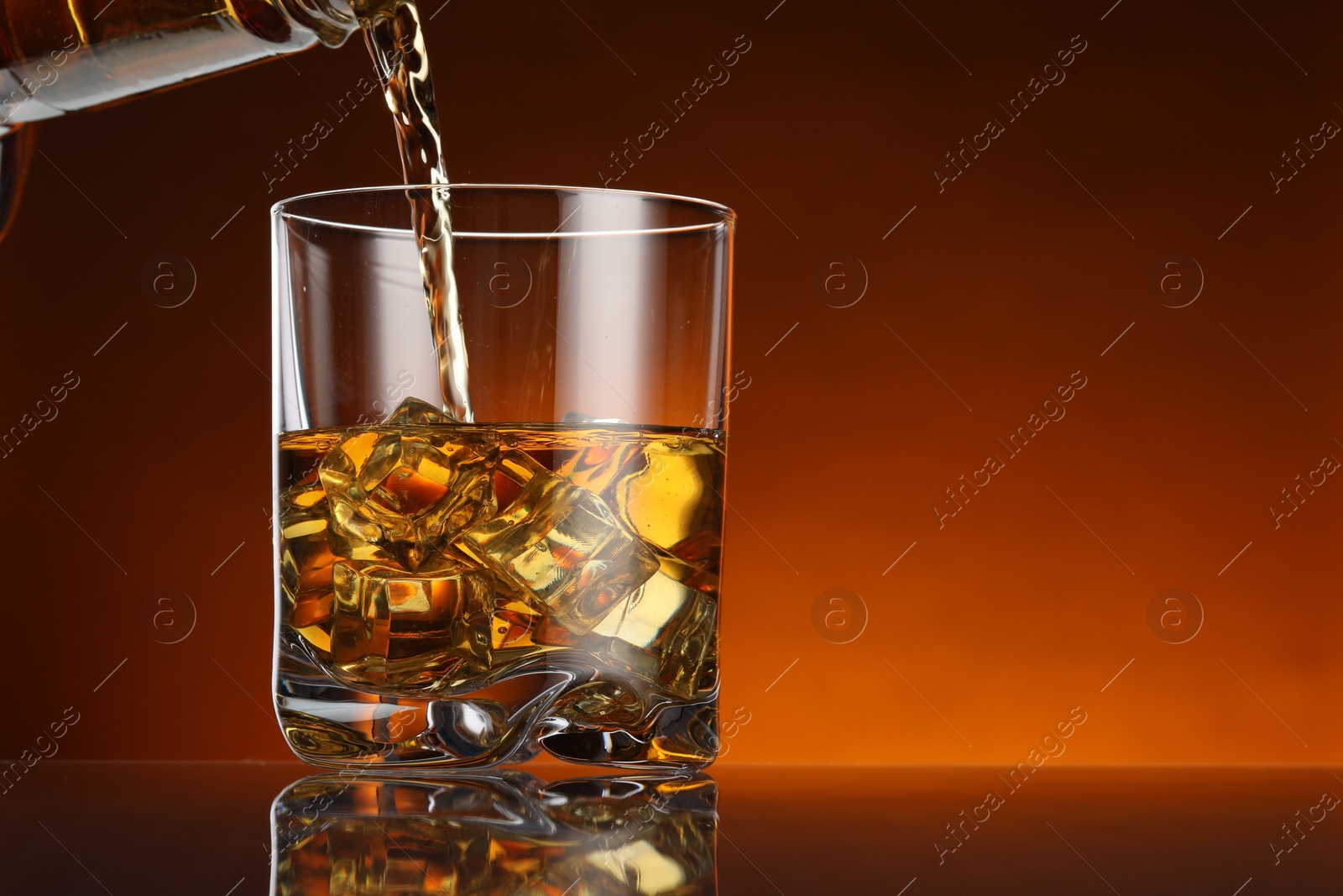 Photo of Pouring whiskey into glass with ice cubes at table against color background, space for text