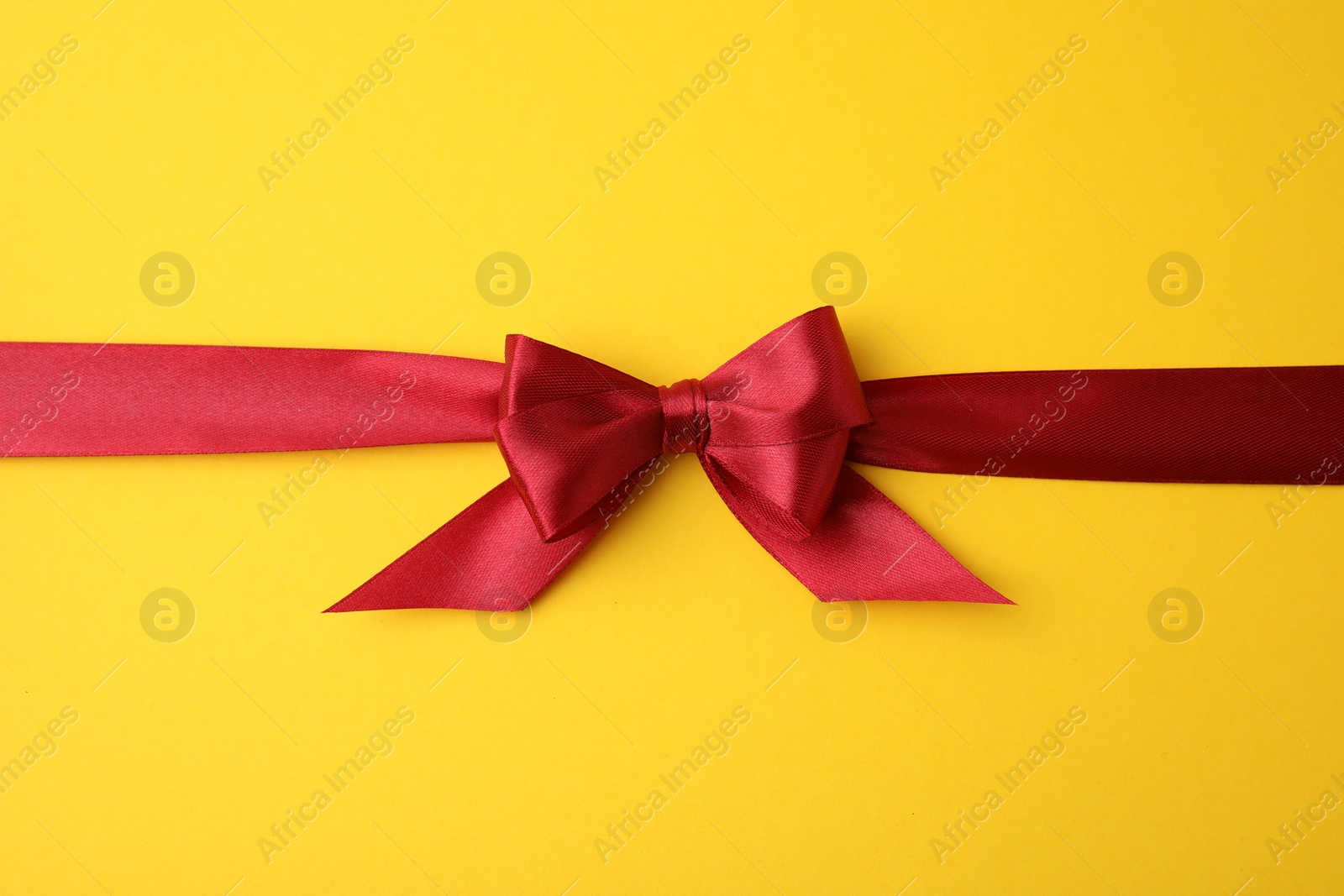 Photo of Red satin ribbon with bow on yellow background, top view