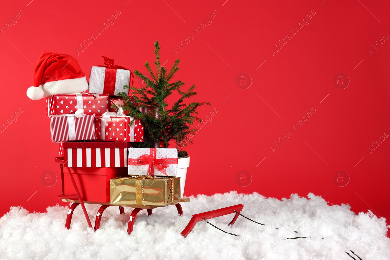 Photo of Sleigh with presents, Santa hat and fir tree in artificial snow on red background. Space for text