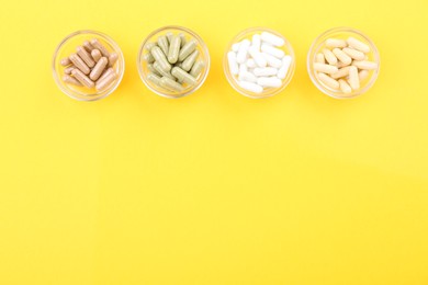 Different vitamin capsules in glass bowls on yellow background, flat lay. Space for text