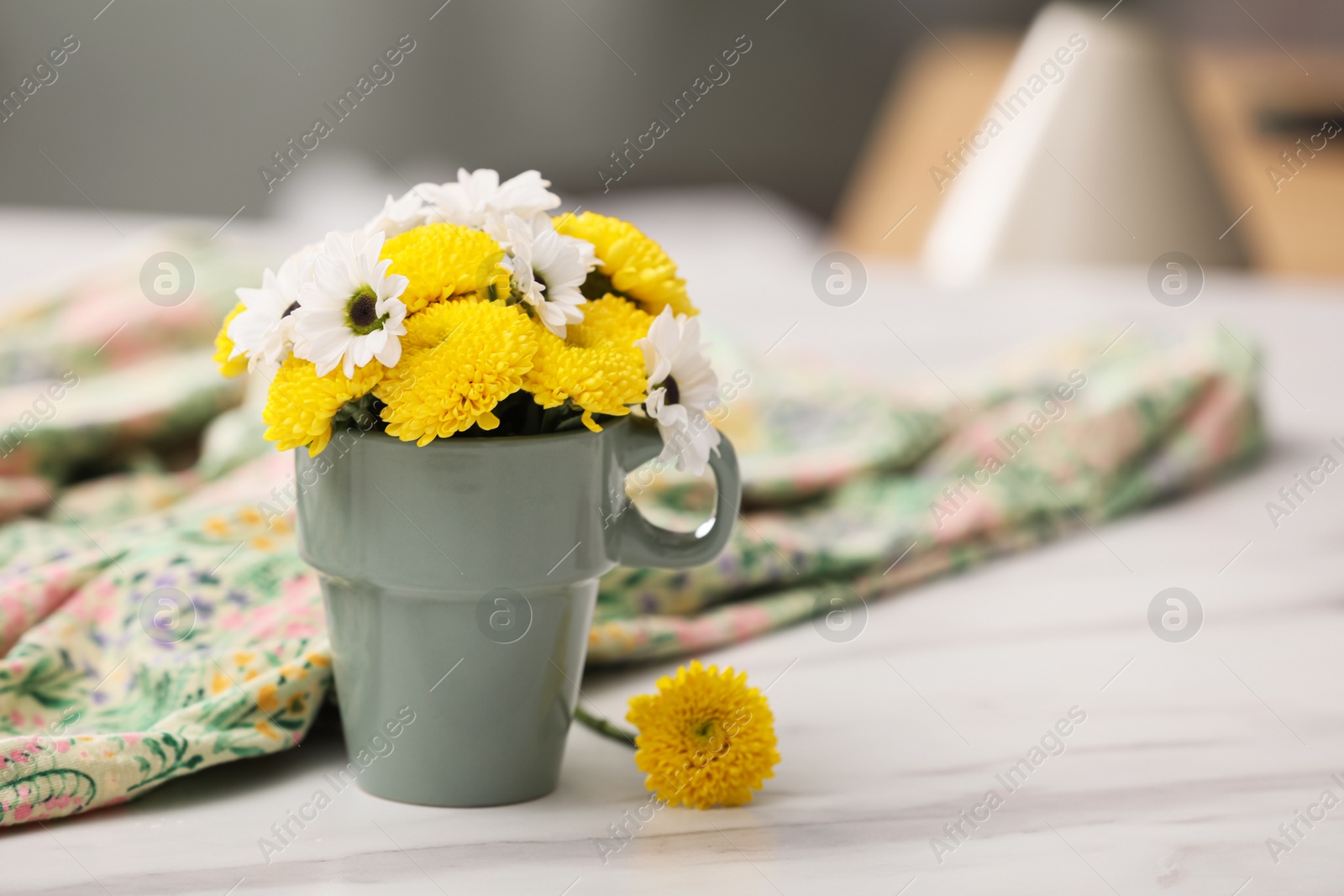Photo of Beautiful bright flowers in cup and fabric on white marble table, space for text
