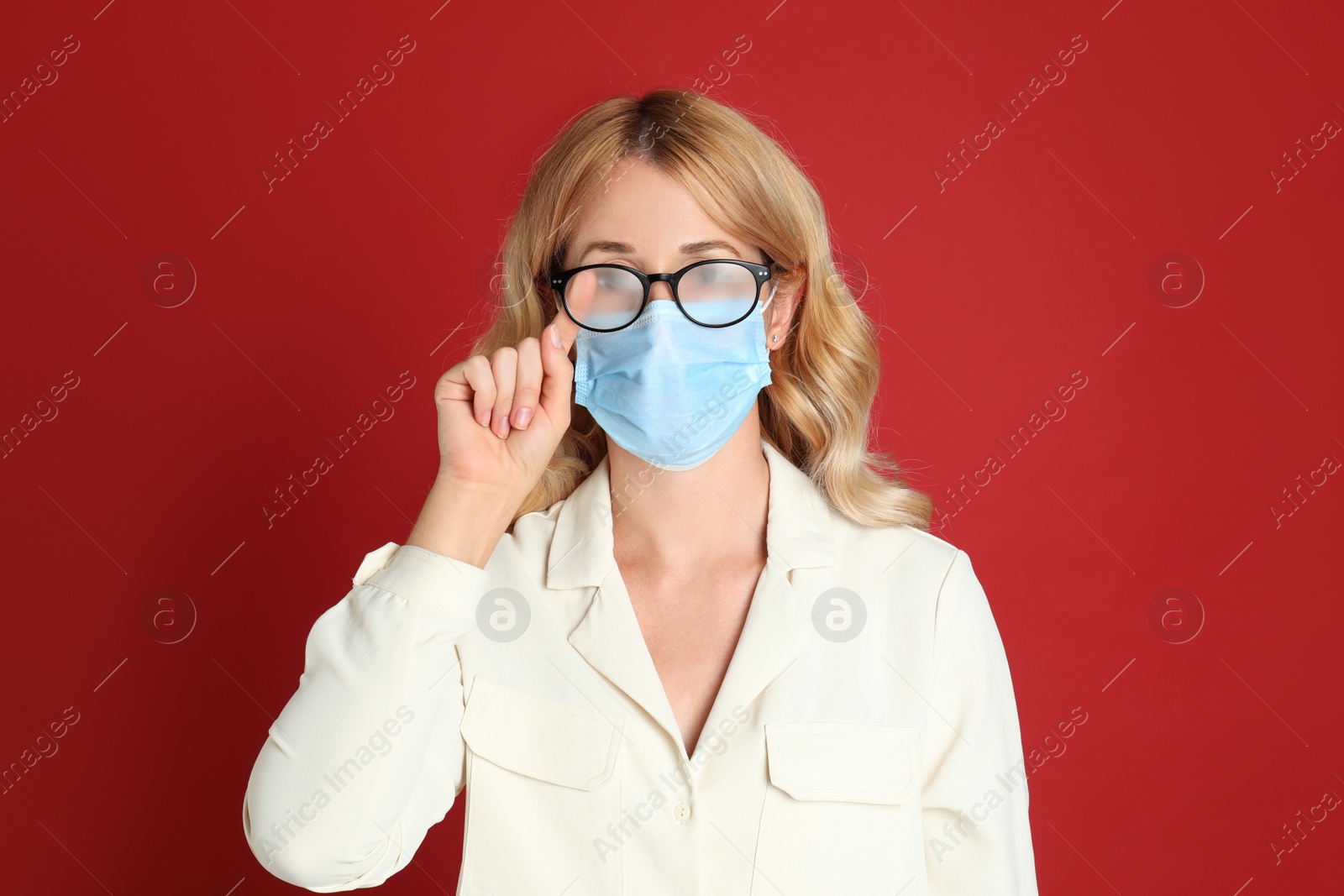 Photo of Woman wiping foggy glasses caused by wearing medical mask on red background