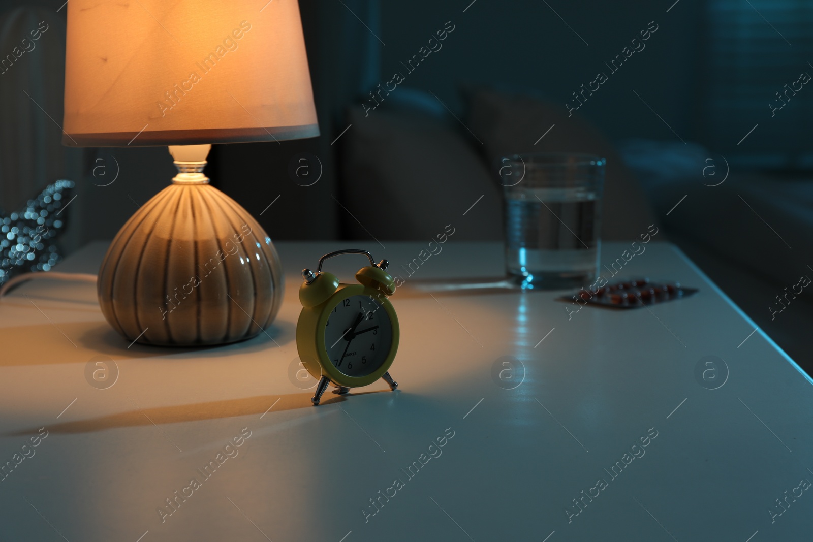 Photo of Insomnia treatment. Glass of water, pills and alarm clock on bedside table in bedroom at night, selective focus