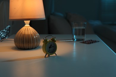 Photo of Insomnia treatment. Glass of water, pills and alarm clock on bedside table in bedroom at night, selective focus