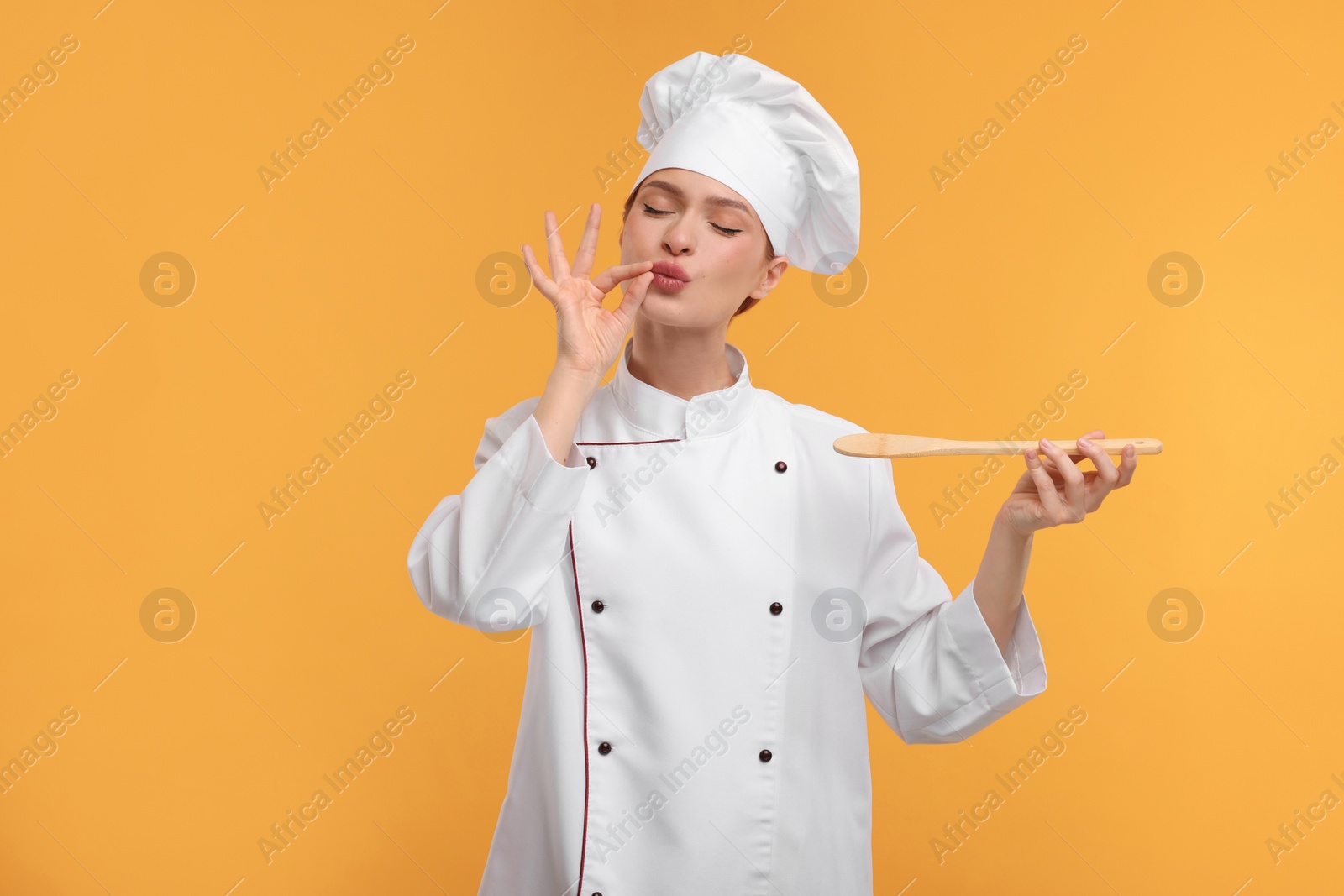 Photo of Young chef in uniform holding wooden spoon and showing perfect sign on orange background