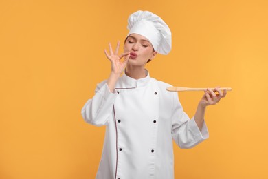 Photo of Young chef in uniform holding wooden spoon and showing perfect sign on orange background