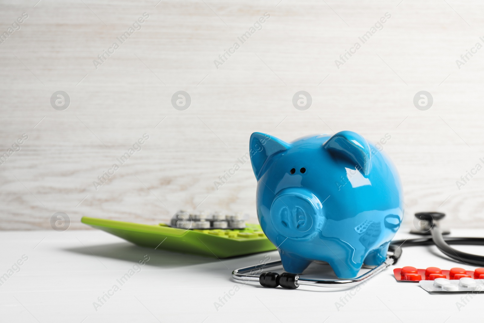 Photo of Light blue ceramic piggy bank, stethoscope and pills on white wooden table, space for text. Medical insurance