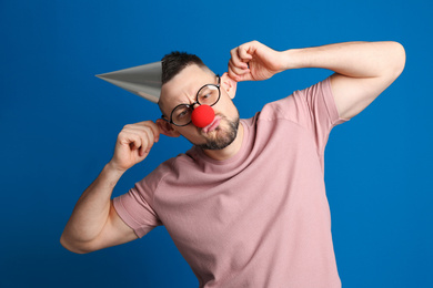 Photo of Funny man with glasses, party hat and clown nose on blue background. April fool's day