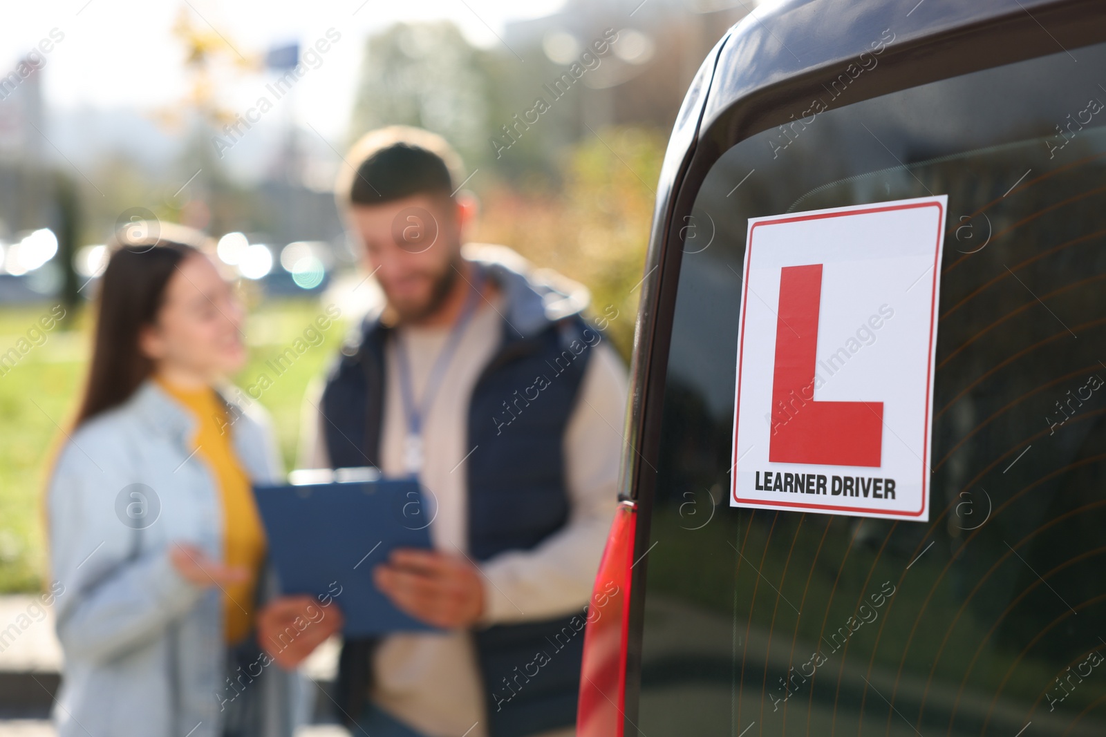 Photo of Learner driver and instructor with clipboard near car outdoors, selective focus on L-plate. Driving school
