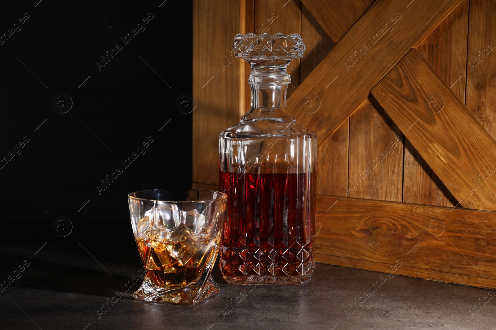 Photo of Whiskey in glass and bottle near wooden crate on dark table against black background