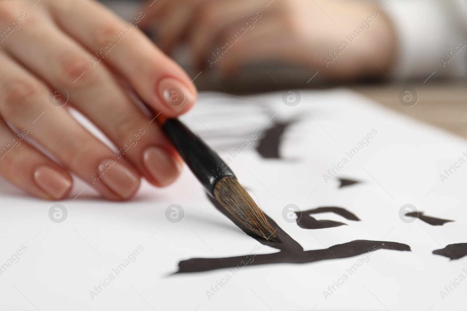 Photo of Calligraphy. Woman with brush writing hieroglyphs on paper at table, closeup