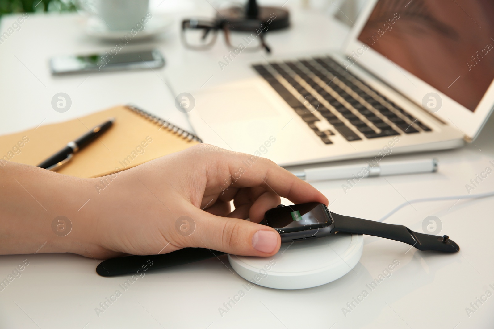 Photo of Man putting smartwatch onto wireless charger at white table, closeup. Modern workplace accessory