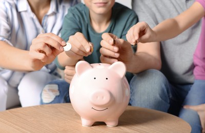 Family putting coins into piggy bank at table indoors, closeup