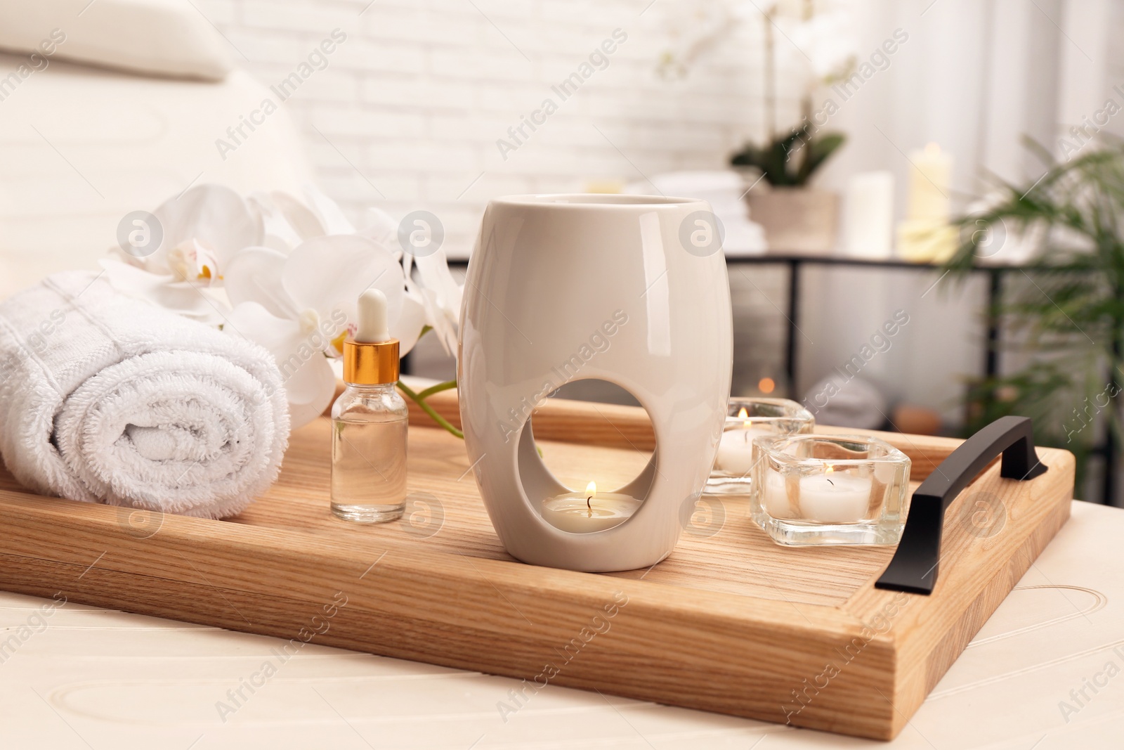 Photo of Aroma lamp, bottle of oil, rolled towel and candles on wooden tray indoors