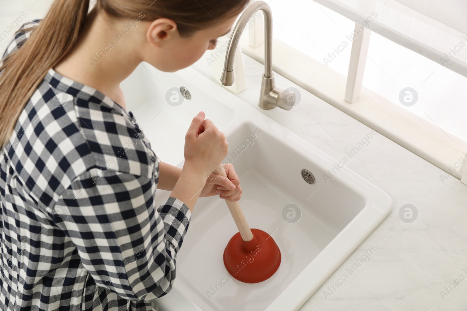 Photo of Young woman using plunger to unclog sink drain in kitchen, above view