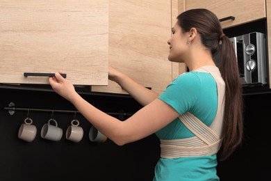 Photo of Woman with orthopedic corset opening cupboard in kitchen