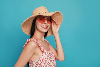 Beautiful young woman with straw hat and heart shaped sunglasses on light blue background. Space for text