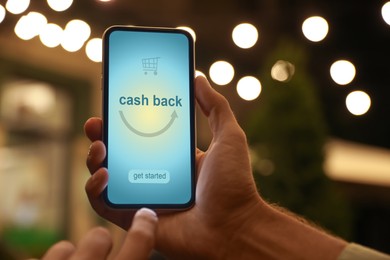 Image of Man using smartphone with word Cashback and shopping cart icon outdoors at night, closeup