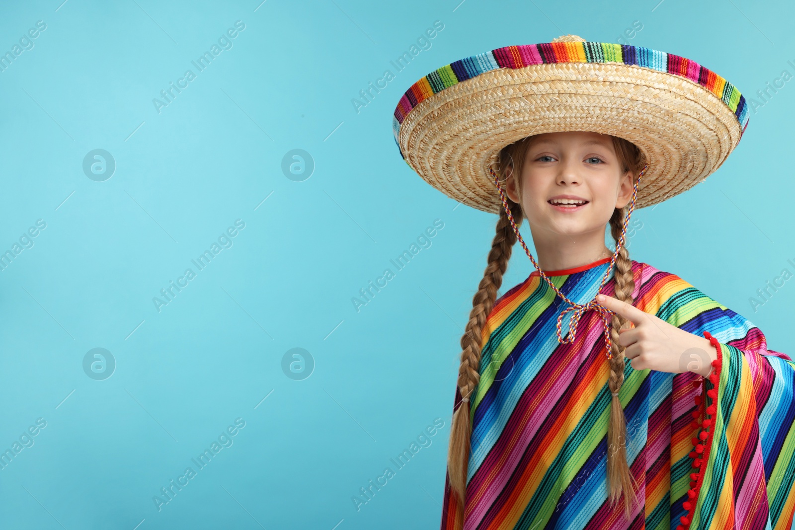 Photo of Cute girl in Mexican sombrero hat and poncho pointing at something on light blue background. Space for text