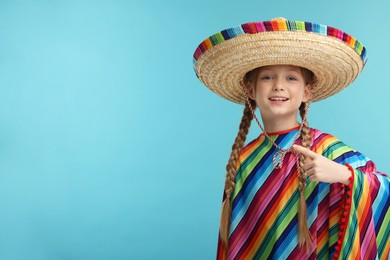 Cute girl in Mexican sombrero hat and poncho pointing at something on light blue background. Space for text