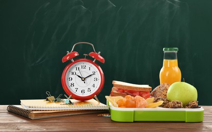 Image of Lunch box with appetizing food and alarm clock on wooden table near green chalkboard