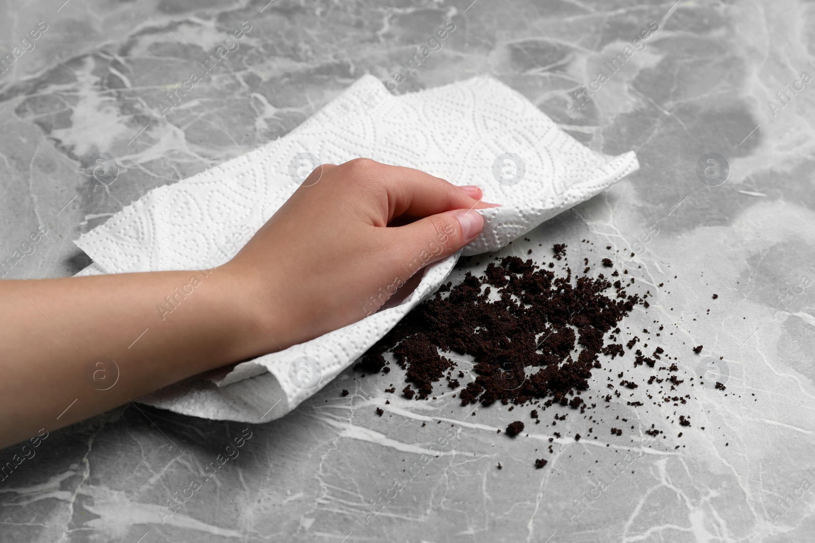 Photo of Woman sweeping scattered coffee grounds with paper towel from marble table, closeup