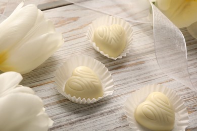 Photo of Delicious heart shaped chocolate candies and beautiful tulips on white wooden table, closeup